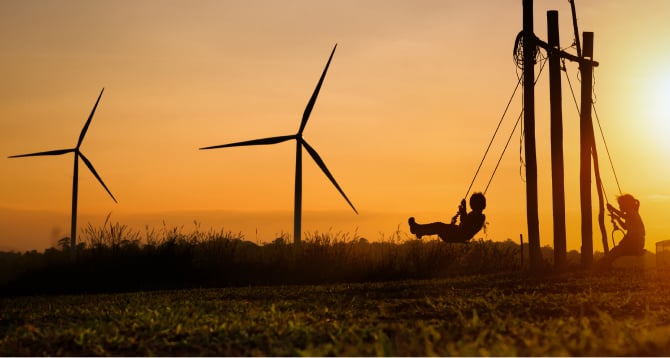 Silhouette of two children outside swinging on a swing set, and two wind turbines in the distance.