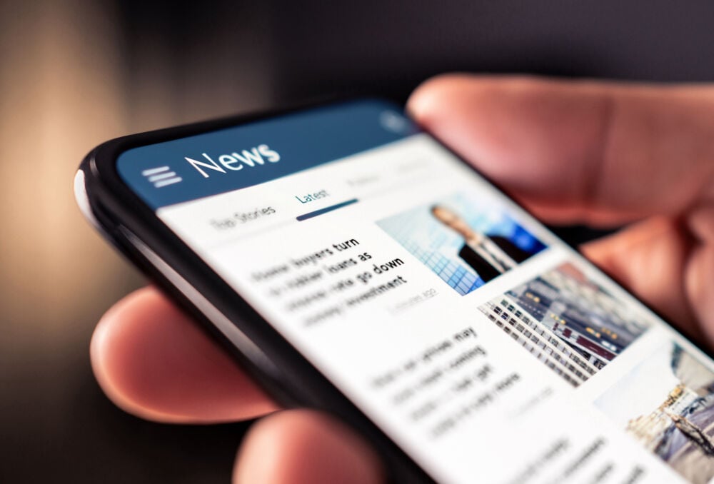 This image shows a close-up of a person holding a smartphone displaying the latest news. The screen features a "News" section with headlines and images, representing digital news consumption. The image captures the ease and immediacy of accessing current events and updates via mobile devices, highlighting the modern way of staying informed